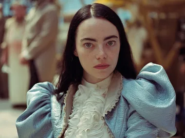 A woman with long dark hair in a blue costume looks seriously at the camera, blurred background with people.