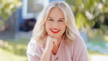 Smiling woman with red lipstick and blonde hair, wearing a pink top, resting her chin on her hand outdoors.