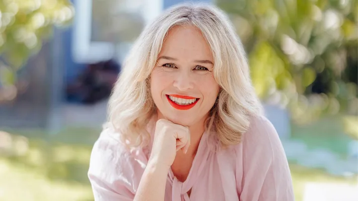 Smiling woman with red lipstick and blonde hair, wearing a pink top, resting her chin on her hand outdoors.