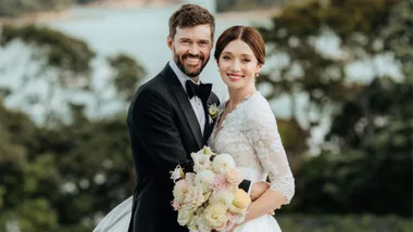 A bride in a lace dress and groom in a tuxedo smile at the camera, holding a bouquet, with greenery in the background.