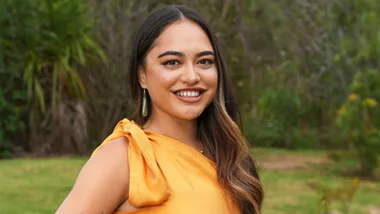 Smiling woman in an orange dress, standing outside with a green leafy background.