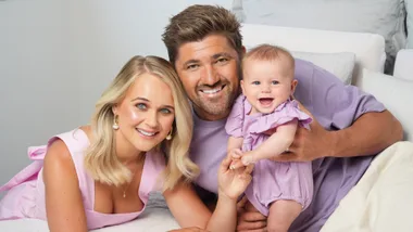 A smiling family with a woman, man, and baby dressed in coordinated purple and pink outfits, lying on a bed.