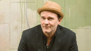 Man in a straw hat and dark blazer against a textured green wall background.