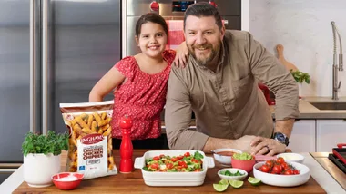 Man and girl smiling in kitchen with ingredients and a pack of Ingham’s Crumbed Chicken Chipees on the counter.