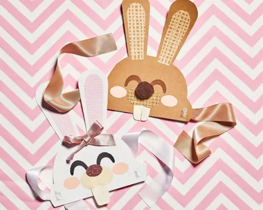 Marbled Easter eggs on a plate beside bunny masks with ribbons on pink zigzag background.