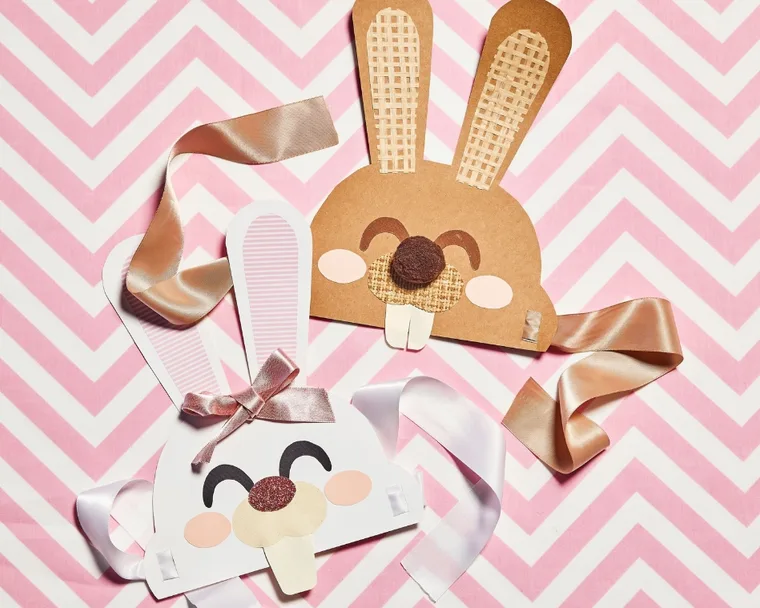 Marbled Easter eggs on a plate beside bunny masks with ribbons on pink zigzag background.