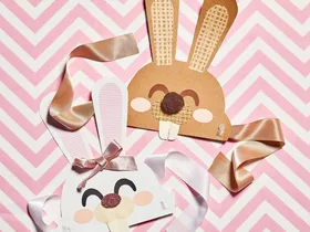 Marbled Easter eggs on a plate beside bunny masks with ribbons on pink zigzag background.