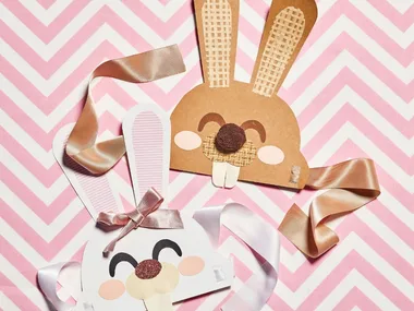 Marbled Easter eggs on a plate beside bunny masks with ribbons on pink zigzag background.