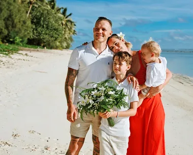 A family posing on a sunny beach; a man, woman, two children, and a boy holding a bouquet of flowers.