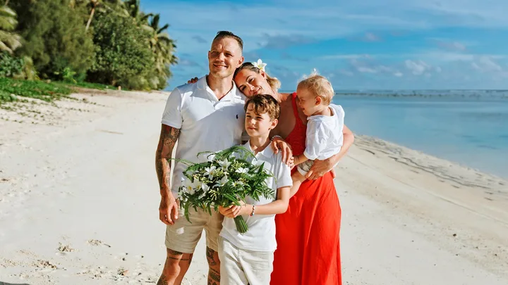 A family posing on a sunny beach; a man, woman, two children, and a boy holding a bouquet of flowers.