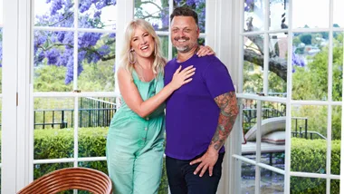 Two people smiling and embracing in a bright room with large windows and blooming purple flowers outside.