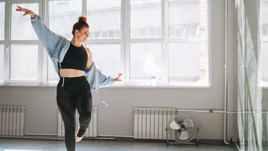 A woman in activewear dances joyfully in a bright studio with large windows.