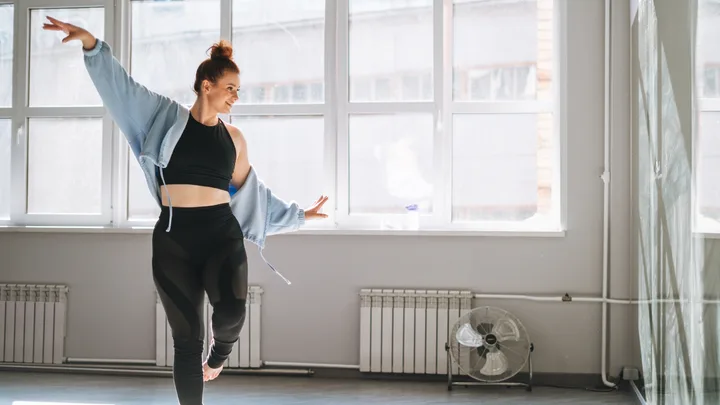 A woman in activewear dances joyfully in a bright studio with large windows.