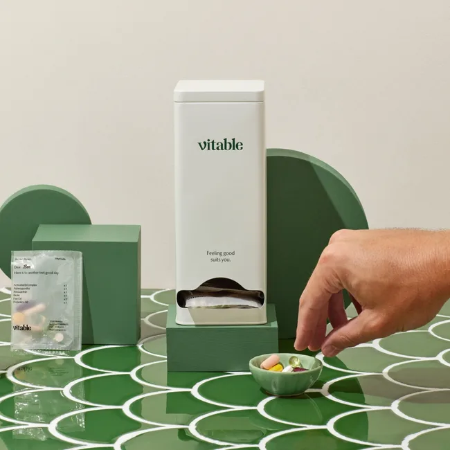 Vitable dispenser on green surface with hand reaching for a bowl of supplements.