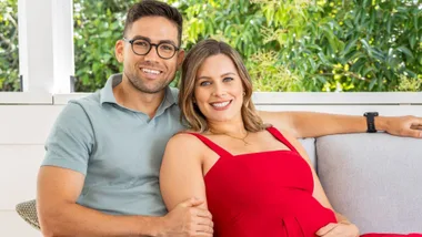 A smiling couple seated together, with a leafy background, woman in red dress, man in light blue shirt.
