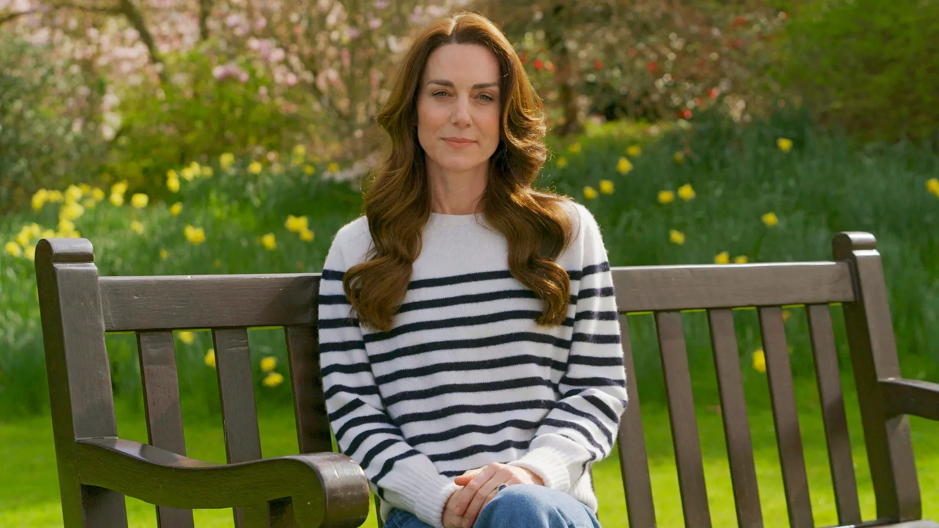 Woman in striped sweater sitting on a bench in a garden with daffodils and greenery in the background.