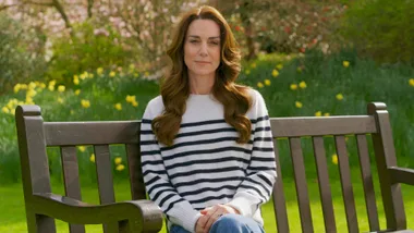 Woman in striped sweater sitting on a bench in a garden with daffodils and greenery in the background.