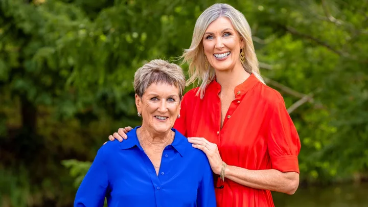Two women smiling; one in a blue shirt and the other in a red dress, standing outdoors with green foliage in the background.