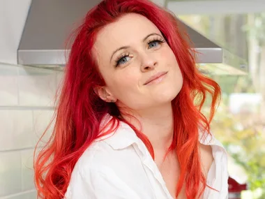 A person with red hair smiling, wearing a white shirt, in a kitchen with a garden view outside.