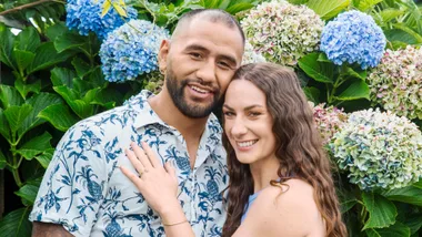 A couple smiling in front of hydrangea flowers, with the woman wearing a blue dress and the man in a patterned shirt.