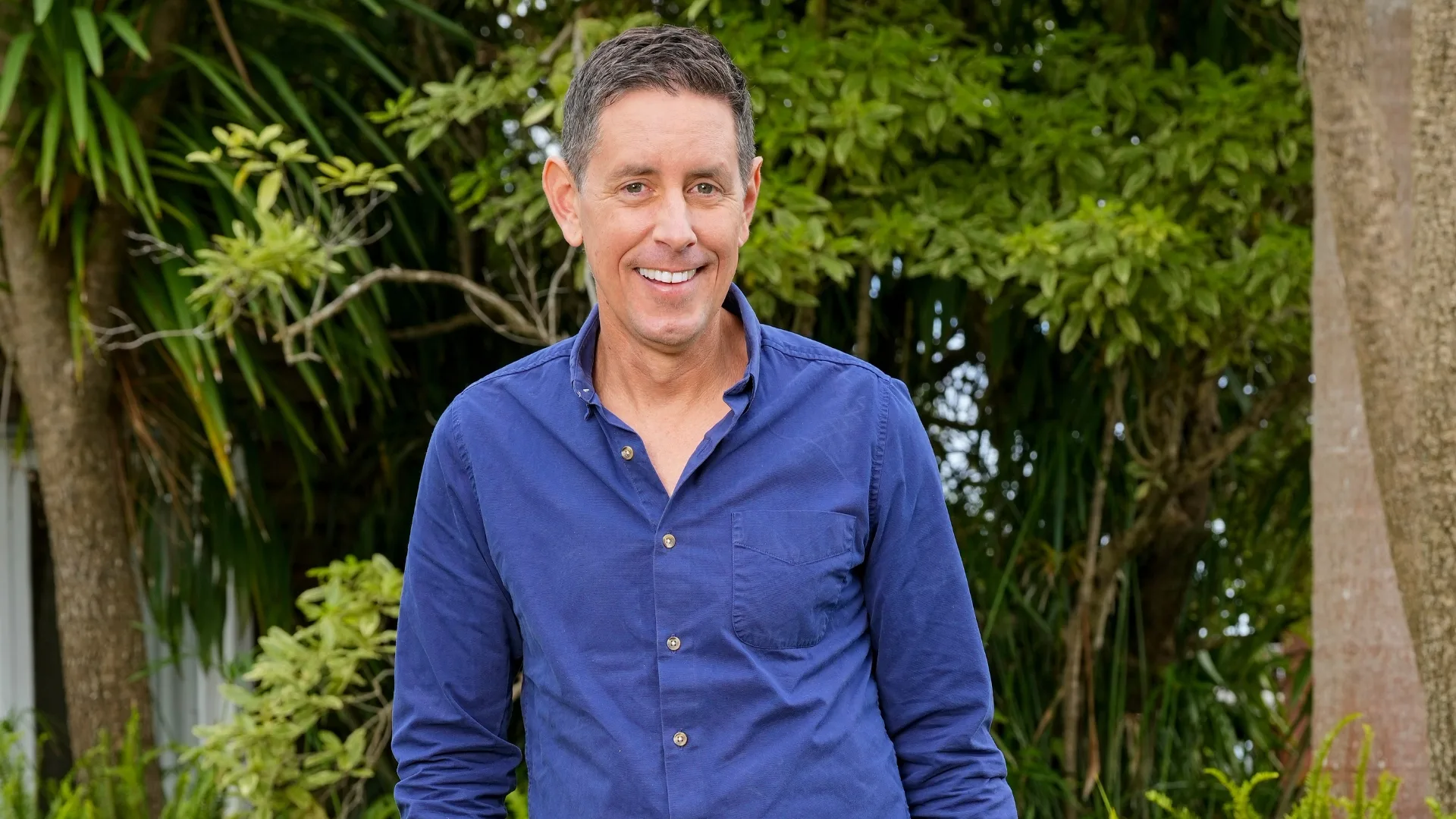 A person in a blue shirt smiles outdoors, surrounded by greenery.