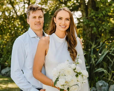 A couple posing outdoors; woman in white dress holds a bouquet, man in a light shirt, greenery in the background.