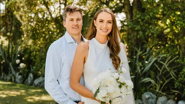 A couple posing outdoors; woman in white dress holds a bouquet, man in a light shirt, greenery in the background.