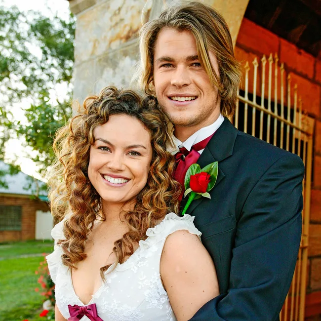 A couple in wedding attire smiles outdoors; the man wears a suit with a red rose boutonniere, and the woman a white dress.