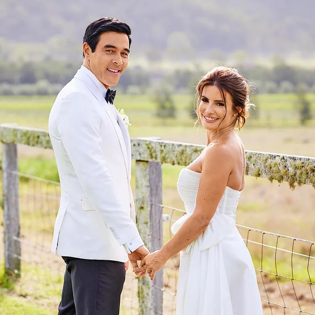 A couple in wedding attire holding hands outdoors; the man in a white suit and woman in a white dress, both smiling.