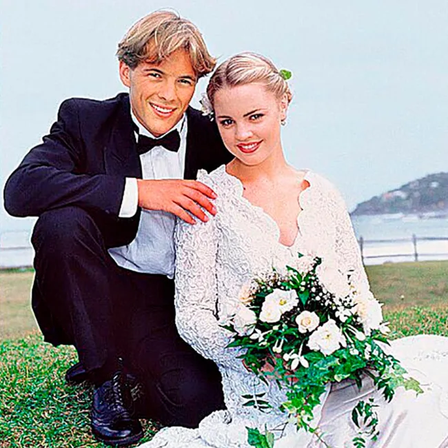 A bride in a white dress holds flowers, with a groom in a black suit kneeling beside her, outdoors near the ocean.