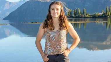 A woman smiling by a serene lake and mountain landscape, wearing a floral top.