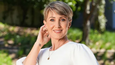 A woman with short hair in a white top smiles outdoors, touching her ear. A star tattoo is visible on her wrist.
