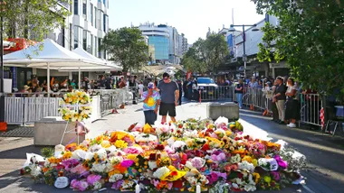 A crowd gathers around a large floral tribute on a city street, with two people standing together in contemplation.
