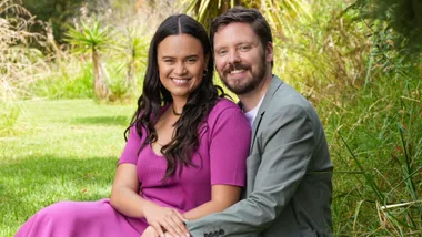Couple sitting on grass, smiling, surrounded by greenery. Woman in pink dress, man in gray blazer.