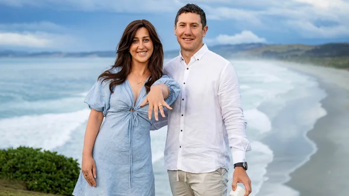 A couple smiling at the camera on a beach; the woman shows her engagement ring.