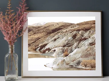 Framed landscape photo of rocky hills above a calm lake, next to pink flowers in a glass vase on a white surface.