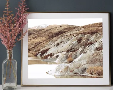 Framed landscape photo of rocky hills above a calm lake, next to pink flowers in a glass vase on a white surface.
