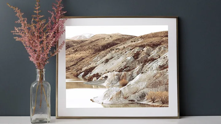 Framed landscape photo of rocky hills above a calm lake, next to pink flowers in a glass vase on a white surface.