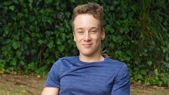 Young man in a blue shirt smiles against a leafy green background.