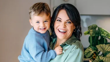 Woman smiling while holding a young child in a blue sweater indoors, with a plant in the background.