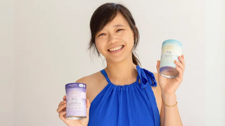 Young woman in a blue dress smiling, holding two cans of Mammas Milk Bar oat milk products against a plain background.