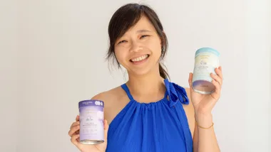Young woman in a blue dress smiling, holding two cans of Mammas Milk Bar oat milk products against a plain background.