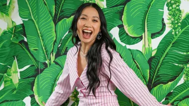 Smiling woman in a pink striped shirt, standing against a background of large green leaves.