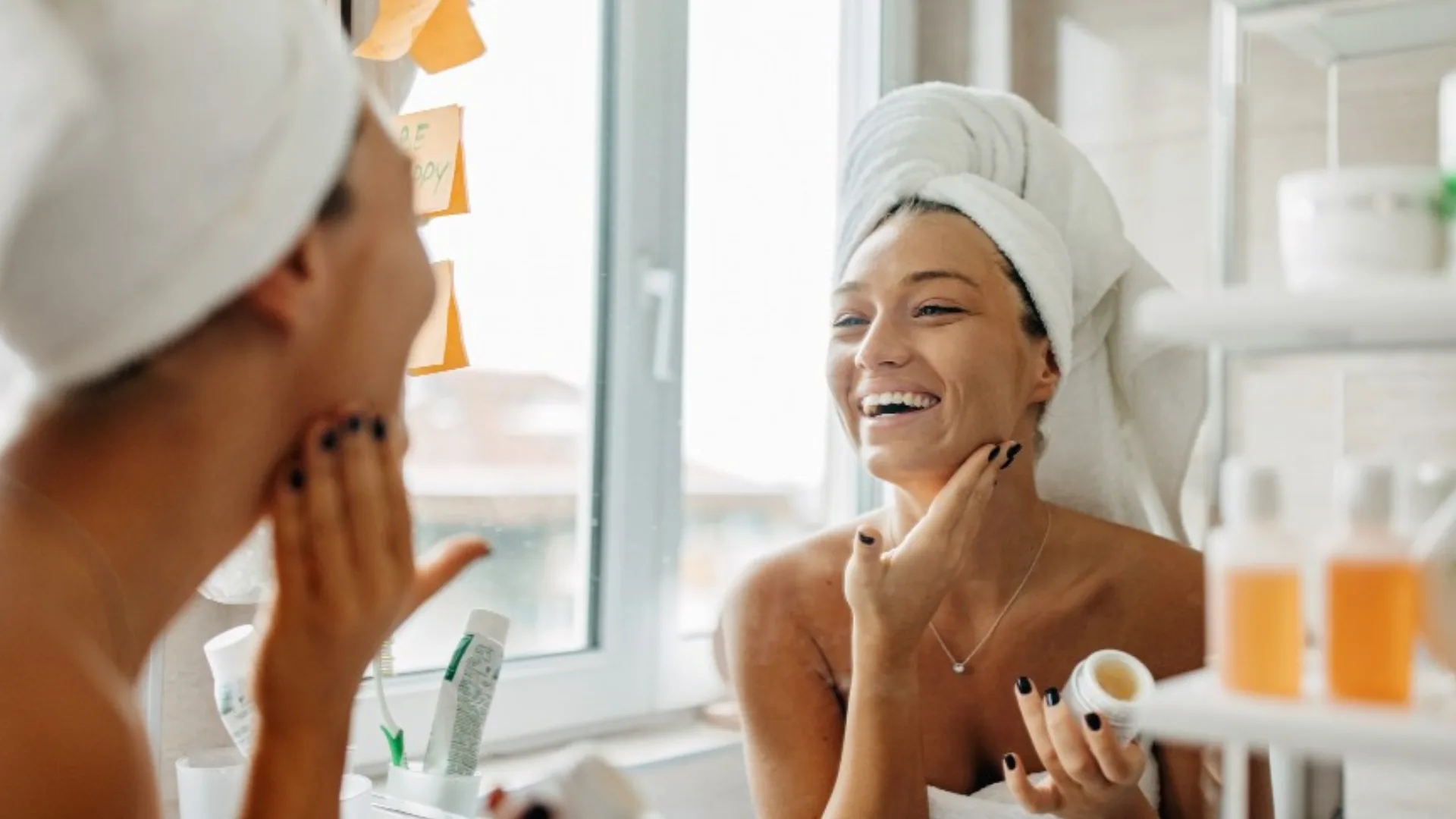 Woman in towel smiles in mirror applying cream; bottles and sticky notes are visible.