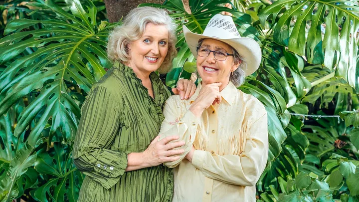 Two elderly women smiling, one in a green blouse and the other in a cowboy hat, standing in front of lush greenery.