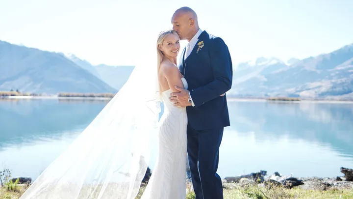 Bride and groom embrace by a serene lake with mountains in the background, groom kisses bride's forehead.
