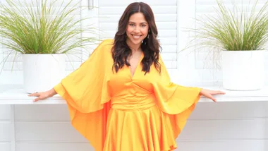 A woman in a bright orange dress smiles in front of potted plants with white shutters in the background.