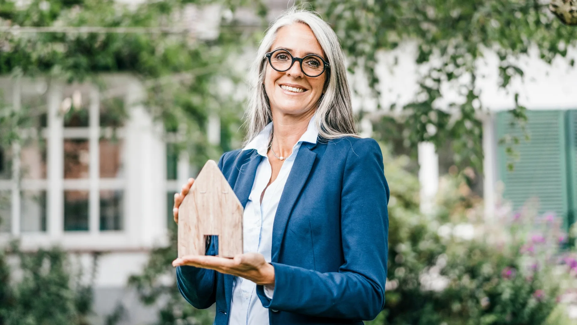 A woman with glasses and gray hair holds a small wooden house model with a garden in the background.
