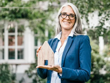 A woman with glasses and gray hair holds a small wooden house model with a garden in the background.