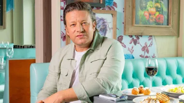 Jamie Oliver sitting off the side of a restaurant booth seat, looking at the camera
